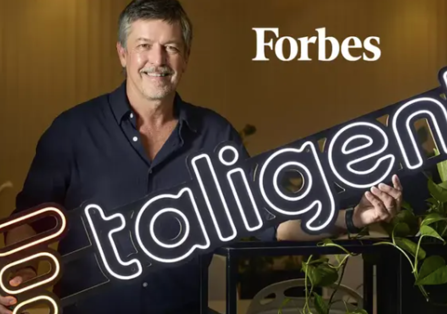 Ejecutivo de Taligent sonriendo junto al logo neón de la marca, con el logo de Forbes de fondo